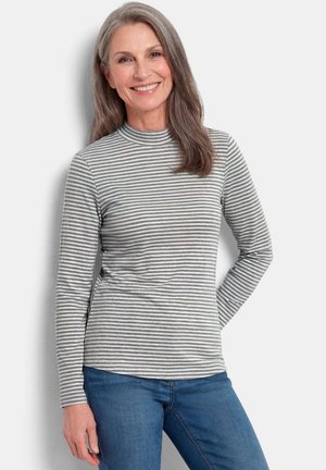 Femme d'âge moyen souriante aux longs cheveux gris portant un t-shirt à manches longues rayé gris et blanc et un jean bleu sur un fond uni.