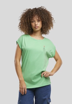 Femme aux cheveux bouclés portant un t-shirt vert clair avec un petit motif main et cœur sur la poitrine, posant avec une main sur la hanche.