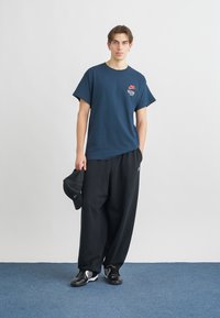 Ναυτικό κοντομάνικο T-shirt με μικρό κόκκινο λογότυπο Nike, σε συνδυασμό με μαύρο παντελόνι σε φαρδιά γραμμή και μαύρα αθλητικά παπούτσια. Κρατάει ένα μαύρο καπέλο.