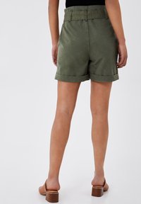 Person, die olivgrüne Shorts mit umgeschlagenem Saum und braune, hochhackige Mules trägt, steht vor einem schlichten hellen Hintergrund, von hinten gezeigt.