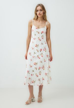 Witte kanten jurk met een roze bloemmotief, spaghettibandjes en een vloeiende silhouet dat tot halverwege de kuit reikt. Casual sandalen maken de look compleet.