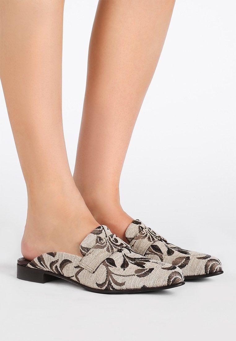 Paire de mocassins beige à enfiler avec un motif floral noir portés pieds nus sur un fond blanc.