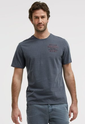 Man with short brown hair wearing a gray striped t-shirt with red text on the chest and gray pants, standing against a plain background.