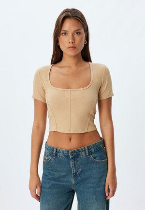 Beige ribbet crop top med firkantet halsudskæring og korte ærmer, med lodret syning og en tætsiddende pasform. Blandet med blå højtaljede jeans.