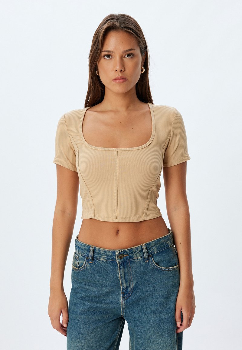 Crop top beige a coste con scollatura quadrata e maniche corte, caratterizzato da cuciture verticali e un design aderente. Abbinato a jeans blu a vita alta.