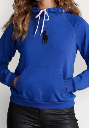 Vrouw draagt een felblauwe hoodie met witte trekkoorden en een zwart polospelerlogo, handen in de voorzak, gecombineerd met zwarte broek.