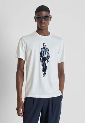 Hombre con gafas de sol oscuras, camiseta blanca con un gráfico de un hombre caminando con chaqueta y pantalones oscuros, y pantalones oscuros, posando frente a un fondo liso.