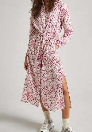 Femme portant une robe midi blanche et rose à motifs avec une fente latérale, manches longues, taille nouée avec pompons, associée à des chaussettes blanches et des baskets.
