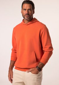 Felpa arancione in morbido tessuto con tasca frontale, polsini a coste e cappuccio. Abbinata a pantaloni chiari e braccialetti in pelle.