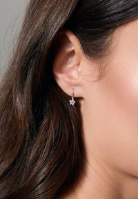 Złote kolczyki w kształcie obrączek z kwiatowym wzorem, z różowymi i białymi akcentami z kamieni szlachetnych, prezentowane obok ucha modelki o długich, ciemnych włosach.