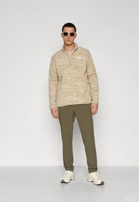 Béžový fleecový pulovr s půlzipem a strukturovaným vzorem, doplněný olivově zelenými kalhotami a bílými teniskami, se zakulacenými bílými slunečními brýlemi.