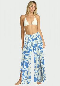 Calças largas de padrão azul e branco com fendas laterais, feitas de tecido leve, combinadas com um top de biquíni branco em forma de triângulo.