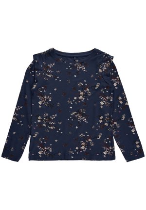 Kind trägt ein blaues, langärmliges, geblümtes Oberteil, bordeauxrote Hose und weiße Sneakers mit lila Akzenten, steht vor weißem Hintergrund.