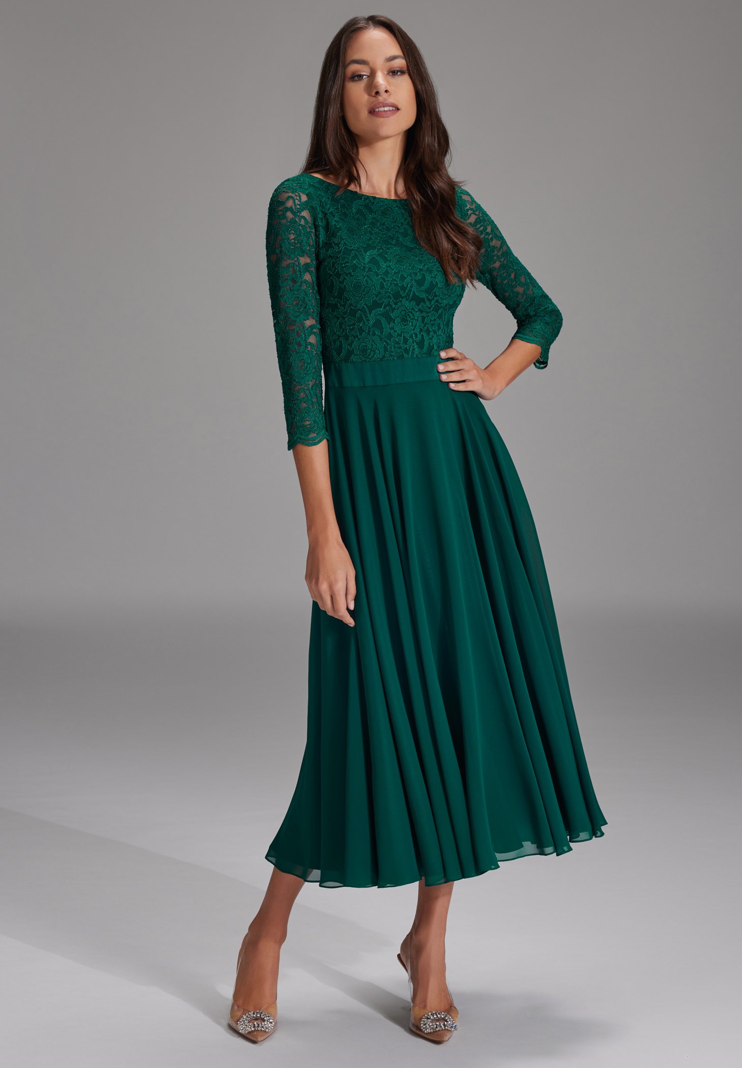 Grønn blonderkjole med trekvart armer og en flytende, plisserte skjørt. Blondekroppen har intrikate mønstre; glatt stoff nedenfor.