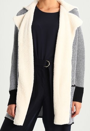 Kardigan z otwartym przodem o teksturowanych, czarno-białych rękawach z dzianiny oraz miękką, białą podszewką z polaru, noszony na czarnej bluzce i spodniach z paskiem w talii.