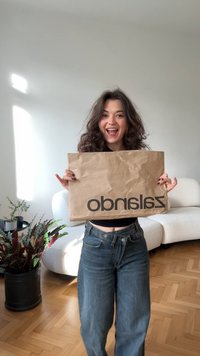 Bolsa de papel marrón con un logo oscuro "zalando" en la parte frontal, sostenida por una persona que lleva jeans azules y una camiseta negra. Fondo liso.