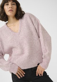 Pulover supradimensionat cu decolteu în V, de culoare roz deschis, realizat dintr-un material moale. Are manșete și tiv cu nervuri, cu un croi lejer și o textură pufoasă.