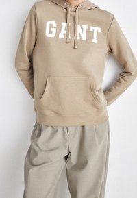 Bežová mikina s kapucí a přední kapsou, s nápisem "GANT" v bílých tučných písmenech. Měkká tkanina, minimalistický design a pohodlný střih.