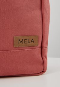 Rosa Canvas-Tasche mit rechteckiger Form, ausgestattet mit einem braunen Lederetikett, das mit "MELA" auf der Seite geprägt ist. Strukturierte Oberfläche.
