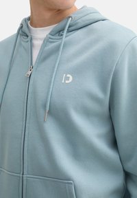 Modrá zateplená mikina na zips vyrobená z jemnej tkaniny. Má predné vrecko, strieborné doplnky a malé logo na hrudi. Kapucňa s šnúrkami.