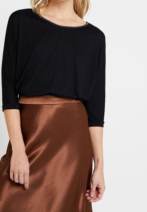 Frau trägt ein lockeres schwarzes Oberteil mit dreiviertel Ärmeln und einen glänzenden braunen Satinsrock, dargestellt vom Hals bis zur Mitte des Oberschenkels.