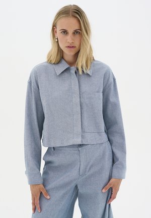 Cropped blue-gray shirt with long sleeves, a collared neckline, and a front pocket. Made from woven fabric with a subtle texture.