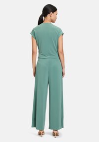 Combinaison verte avec manches courtes, design à jambes larges et texture lisse. Dotée d'une fermeture éclair dans le dos et d'un style minimaliste. Les talons sont beiges.