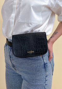 Czarna skórzana torba na pasek o fakturze krokodylej. Posiada zaokrągloną klapę i złote logo. Noszona w talii na jasnych dżinsach.