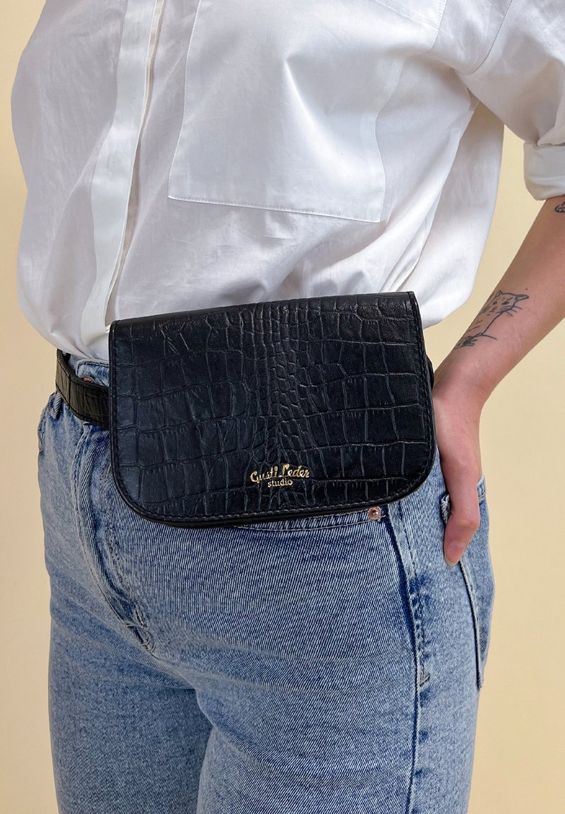 Czarna skórzana torba na pasek o fakturze krokodylej. Posiada zaokrągloną klapę i złote logo. Noszona w talii na jasnych dżinsach.