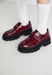 Burgundy lackläderplattformsskor med svart kraftig sula, dubbla remmar med spänne och hjärtformad detalj.