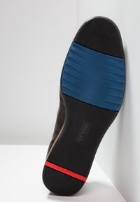 Semelle de chaussure en cuir marron avec une surface noire texturée, dotée d'une bande d'accent bleue et rouge, et d'un logo de marque embossé.