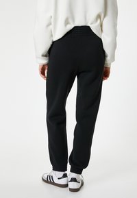 Schwarze Jogginghosen mit einem elastischen Bund und Knöchelrsfen, Strukturstoff, kombiniert mit weißen Sneakers mit schwarzen Streifen.