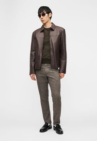 Άνδρας που φοράει σκούρο καφέ δερμάτινο μπουφάν, ελιάς πράσινο πουλόβερ, γκρι παντελόνι tweed, μαύρα loafers και μαύρα γυαλιά ηλίου, στέκεται μπροστά σε λευκό φόντο.