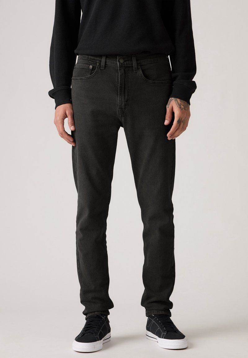 Homme portant un jean noir slim, une chemise noire à manches longues et des baskets noires à semelles blanches, debout devant un fond neutre.