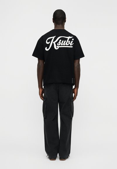 Μαύρο t-shirt Ksubi με μεγάλο λευκό λογότυπο στην πλάτη, κοντά μανίκια, στρογγυλή λαιμόκοψη και ελαφρώς φαρδιά γραμμή. Φορεμένο με μαύρο παντελόνι cargo.