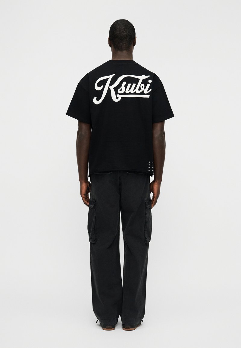 Μαύρο t-shirt Ksubi με μεγάλο λευκό λογότυπο στην πλάτη, κοντά μανίκια, στρογγυλή λαιμόκοψη και ελαφρώς φαρδιά γραμμή. Φορεμένο με μαύρο παντελόνι cargo.