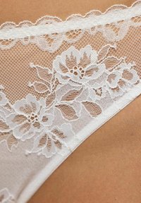 Białe koronkowe bielizna z kwiatowymi wzorami, misternym szyciem i falbaniastym wykończeniem. Materiał ma przejrzystą, delikatną teksturę.