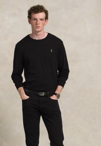 Jovem com cabelo encaracolado a usar um traje completamente preto e um relógio, de pé com as mãos nos bolsos contra um fundo beige liso.