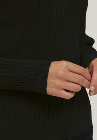 Schwarzer gerippter Pullover mit langen Ärmeln. Hat eine taillierte Form und detaillierte Texturen an den Ärmeln. Hände, die den Stoff anpassen, sind sichtbar.