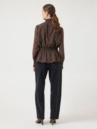 Blouse marron à carreaux avec manches longues, col montant et taille smockée, associée à un pantalon noir ample et des chaussures à talons marron.