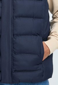 Marineblauer Steppweste mit einem gesteppten Design und horizontalen Nähten, seitlichen Taschen und einer glatten Textur, kombiniert mit einem hellen Creme-Pullover.