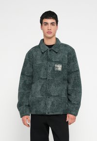 Πράσινο, υφασμάτινο μπουφάν fleece με γιακά, κουμπωτό κλείσιμο και μια τσέπη με κέντημα που διαθέτει γραφικό σχέδιο στο αριστερό στήθος.