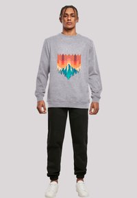 Grauer Sweatshirt mit einem bunten Berg- und Sonnenuntergangsdesign, kombiniert mit schwarzen Jogginghosen und weißen Turnschuhen. Lässiger, bequemer Schnitt.