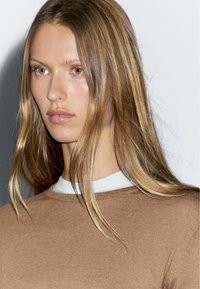 Brązowy sweter robiony na drutach z białym kołnierzykiem, z okrągłym dekoltem i gładką strukturą. Modelka ma długie, falowane włosy.