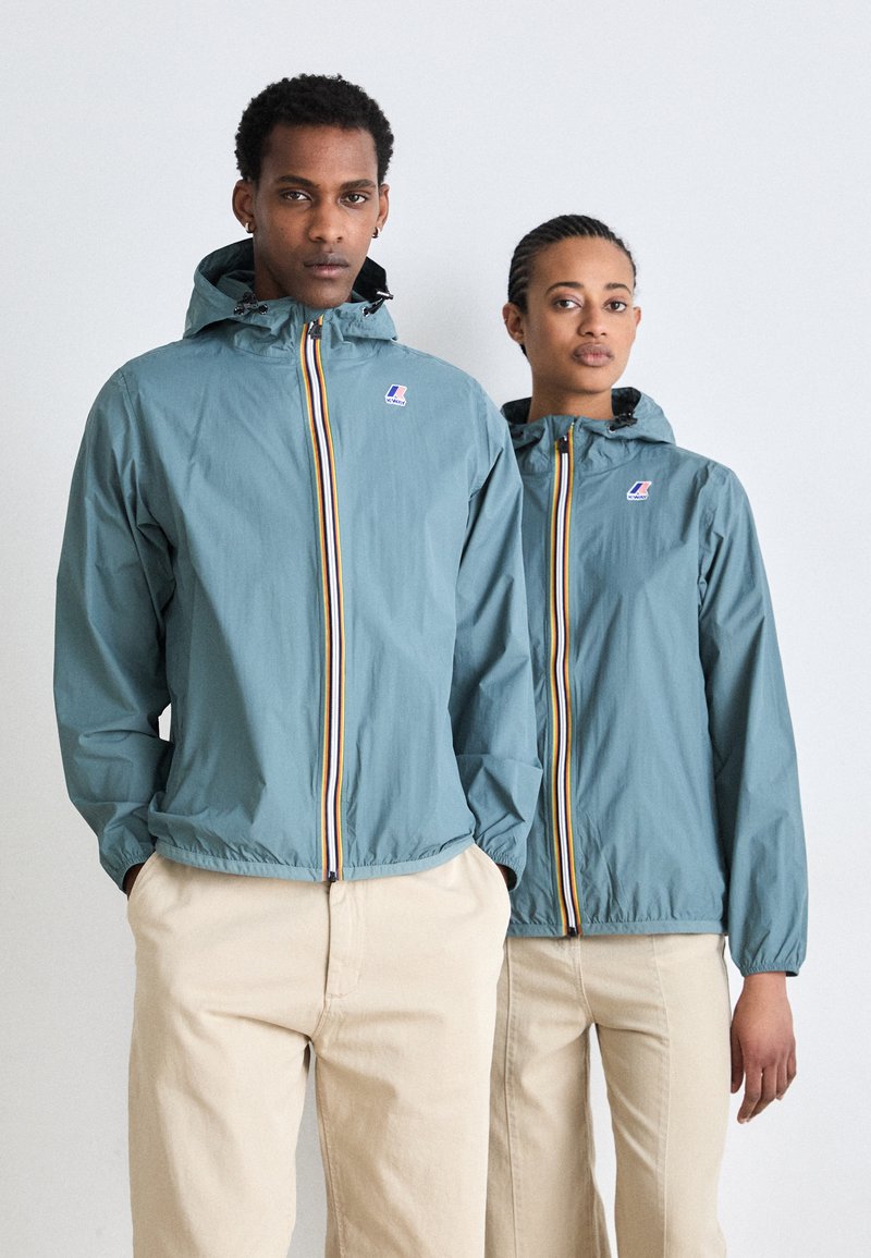 Chaqueta cortavientos azul claro con capucha, que presenta una cremallera frontal con tiras multicolores y un parche con logotipo. Combinada con pantalones beiges.