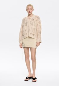 Cardigan beige a maglia aperta con tasche frontali, indossato sopra una gonna strutturata color crema. Sandali neri con dettagli floreali completano l'outfit.