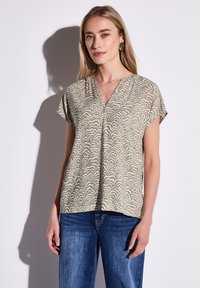 Kortærmet V-hals top i lys beige med et svulmende creme mønster. Glat stof, afslappet pasform, parret med blå jeans.