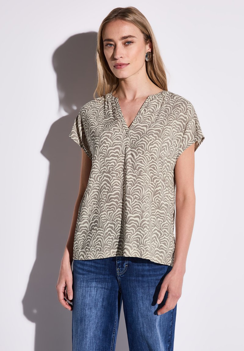 Kortærmet V-hals top i lys beige med et svulmende creme mønster. Glat stof, afslappet pasform, parret med blå jeans.
