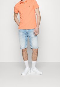 Oranžové polo tričko se třemi knoflíky, světle modré džíny šortky s ohrnutými manžetami, bílé tenisky a bílé ponožky až po kotníky.