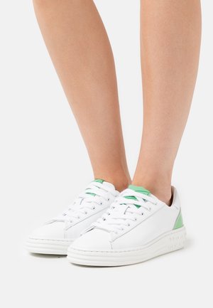 Witte sneakers met groene accenten op de hiel en tong, gedragen op blote voeten tegen een effen lichte achtergrond.